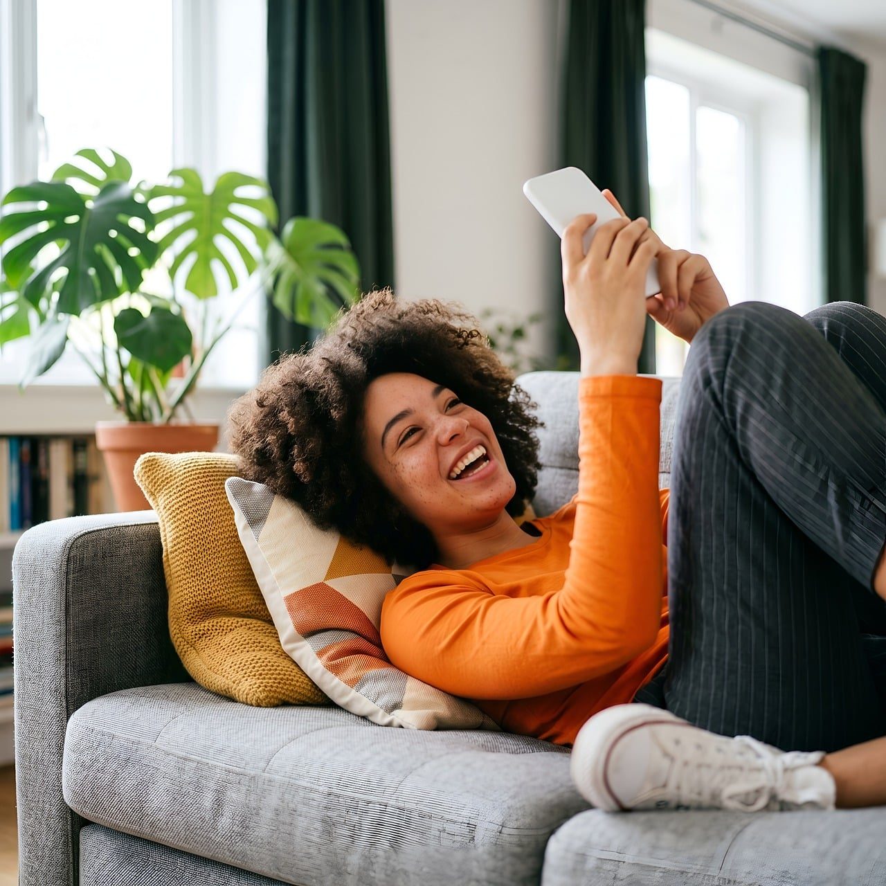 Leende kvinna som ligger i soffan hemma och tittar i mobilen